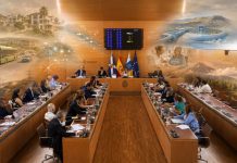Pleno Tenerife mayo 2026: una isla entre gestión y relato Vista fotorrealista del salón de plenos del Cabildo de Tenerife, con los consejeros reunidos y, sobre la escena, imágenes difuminadas que evocan los grandes temas tratados: vivienda, movilidad, agua, energía, servicios sociales, sostenibilidad y protección del territorio.