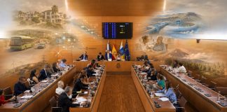 Pleno Tenerife mayo 2026: una isla entre gestión y relato Vista fotorrealista del salón de plenos del Cabildo de Tenerife, con los consejeros reunidos y, sobre la escena, imágenes difuminadas que evocan los grandes temas tratados: vivienda, movilidad, agua, energía, servicios sociales, sostenibilidad y protección del territorio.