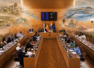 Pleno Tenerife mayo 2026: una isla entre gestión y relato Vista fotorrealista del salón de plenos del Cabildo de Tenerife, con los consejeros reunidos y, sobre la escena, imágenes difuminadas que evocan los grandes temas tratados: vivienda, movilidad, agua, energía, servicios sociales, sostenibilidad y protección del territorio.