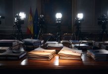 Causas judiciales: el foco selectivo de la indignación mediática Mesa institucional con expedientes judiciales iluminados por focos de prensa, cámaras y micrófonos al fondo, simbolizando la atención mediática selectiva sobre determinadas causas.