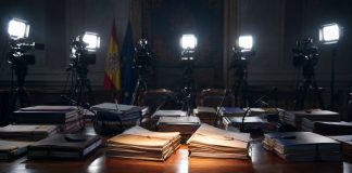 Causas judiciales: el foco selectivo de la indignación mediática Mesa institucional con expedientes judiciales iluminados por focos de prensa, cámaras y micrófonos al fondo, simbolizando la atención mediática selectiva sobre determinadas causas.