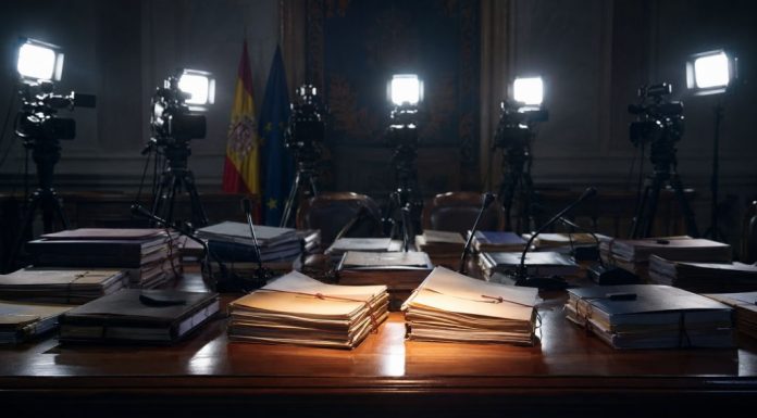 Causas judiciales: el foco selectivo de la indignación mediática Mesa institucional con expedientes judiciales iluminados por focos de prensa, cámaras y micrófonos al fondo, simbolizando la atención mediática selectiva sobre determinadas causas.