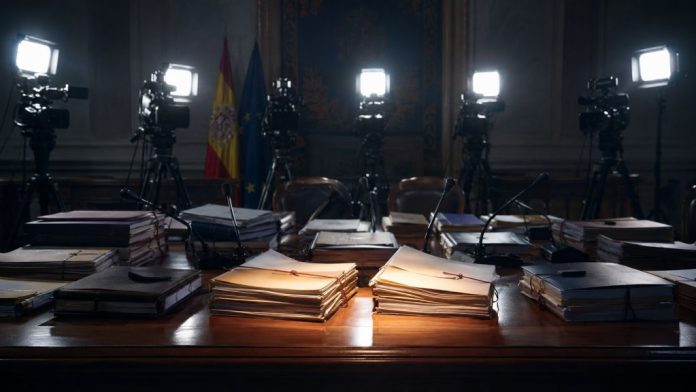 Mesa institucional con expedientes judiciales iluminados por focos de prensa, cámaras y micrófonos al fondo, simbolizando la atención mediática selectiva sobre determinadas causas.