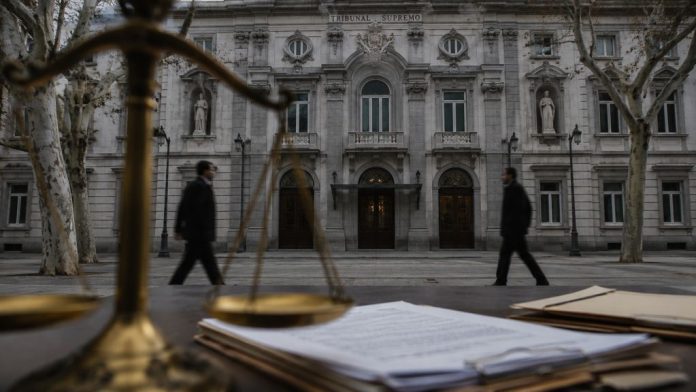 Fachada del Tribunal Supremo con una balanza de justicia desenfocada, documentos judiciales en primer plano y dos figuras anónimas caminando frente al edificio.