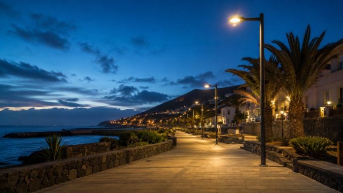 Paseo costero de Candelaria al anochecer, iluminado con luminarias LED de luz cálida junto al mar y con palmeras al fondo.