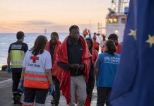 Pacto migratorio: Canarias no puede ser frontera y soledad Personas migrantes son atendidas en un muelle canario al amanecer por personal humanitario y servicios públicos, con una bandera europea desenfocada al fondo que simboliza la responsabilidad compartida de Europa ante la migración.