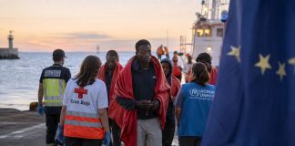 Pacto migratorio: Canarias no puede ser frontera y soledad Personas migrantes son atendidas en un muelle canario al amanecer por personal humanitario y servicios públicos, con una bandera europea desenfocada al fondo que simboliza la responsabilidad compartida de Europa ante la migración.
