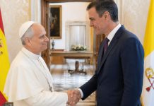 Papa y Sánchez: una sintonía que incomoda El papa León XIV y Pedro Sánchez se saludan cordialmente en una sala institucional, con las banderas de España y la Santa Sede al fondo, en una escena que representa diálogo diplomático y buena sintonía.