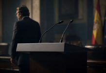 Aznar y el PP: la brújula de la crispación Atril institucional vacío con micrófonos en primer plano y la silueta desenfocada de un expresidente alejándose en una sala oficial, símbolo de una influencia política persistente.