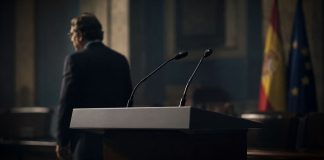 Aznar y el PP: la brújula de la crispación Atril institucional vacío con micrófonos en primer plano y la silueta desenfocada de un expresidente alejándose en una sala oficial, símbolo de una influencia política persistente.