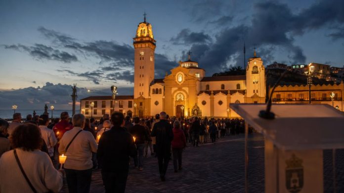 Procesión de las candelas en la plaza de la Basílica de Candelaria al anochecer, con un atril institucional desenfocado en primer plano.