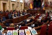 Pleno del Parlamento: sanidad, leyes y confrontación Vista panorámica del pleno del Parlamento de Canarias durante una sesión, con diputados en el hemiciclo y documentos sobre sanidad, ciencia, migración y turismo.