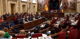 Pleno del Parlamento: sanidad, leyes y confrontación Vista panorámica del pleno del Parlamento de Canarias durante una sesión, con diputados en el hemiciclo y documentos sobre sanidad, ciencia, migración y turismo.