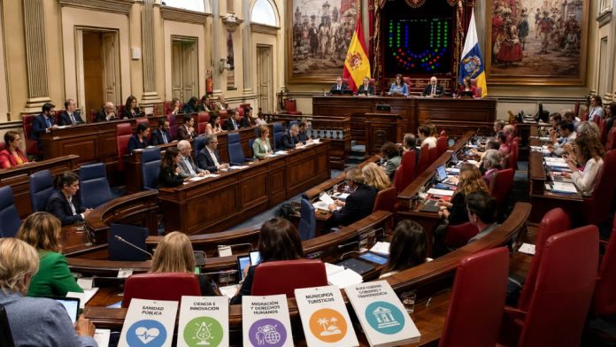 Clavijo 1 Vista panorámica del pleno del Parlamento de Canarias durante una sesión, con diputados en el hemiciclo y documentos sobre sanidad, ciencia, migración y turismo.