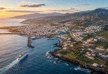 Europa y Canarias: la estrategia que debe cambiar la insularidad Vista aérea al atardecer de una localidad costera canaria, con un ferry saliendo del puerto, zonas agrícolas, viviendas y aerogeneradores en las laderas.