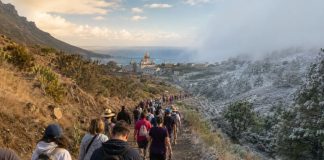 Peregrinación en febrero: Candelaria ante el cambio climático Peregrinos caminan hacia Candelaria por un sendero de Tenerife entre un paisaje seco de verano y otro más frío, simbolizando la adaptación de la tradición al cambio climático.