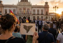 Apoyo a Ceuta: ¿solidaridad o utilización política? Ciudadanos concentrados ante un ayuntamiento con banderas españolas mientras una mujer sostiene una silueta de Ceuta y los medios cubren el acto.