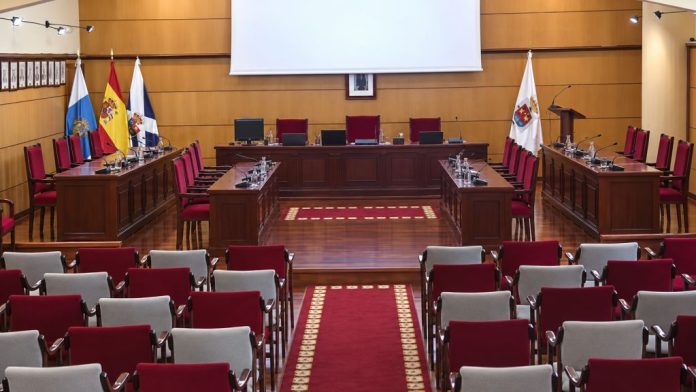 Sala de plenos del Ayuntamiento de Candelaria vacía, con las mesas de los concejales, la presidencia, las banderas institucionales y las butacas del público.