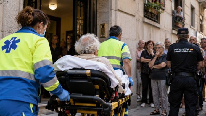 Mujer mayor sale en camilla de un edificio de Madrid acompañada por sanitarios, con vecinos y Policía Nacional durante un desahucio.