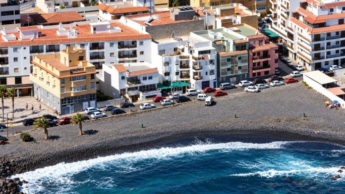 Vista aérea de la playa de Olegario, en Candelaria, con su arena volcánica negra, la calle La Piscina, los edificios del entorno y el océano Atlántico.